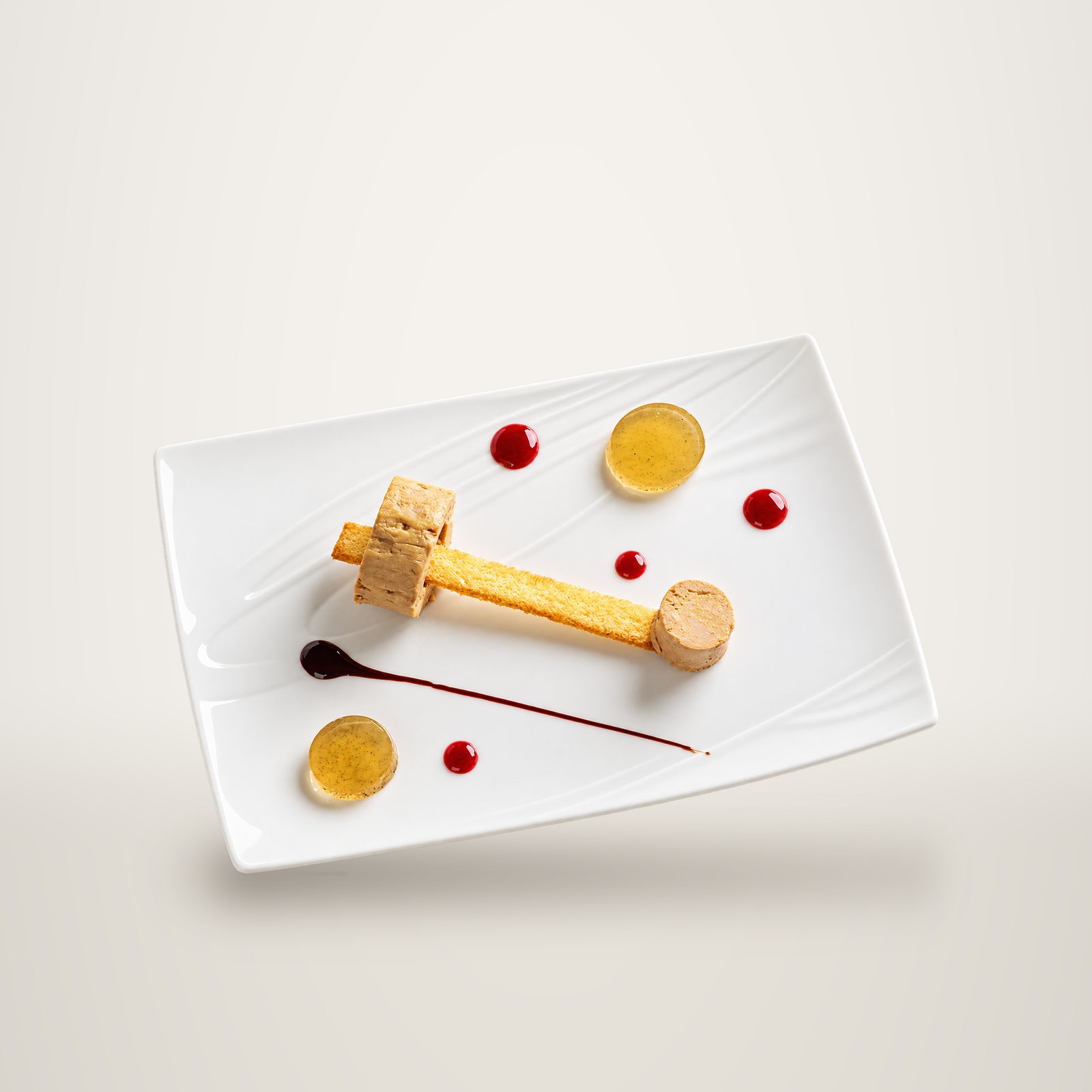 Vue de dessus d'une assiette blanche rectangulaire avec du foie gras sur un bâtonnet de brioche, des disques de gelée, des points de coulis rouge et un trait de sauce sombre.