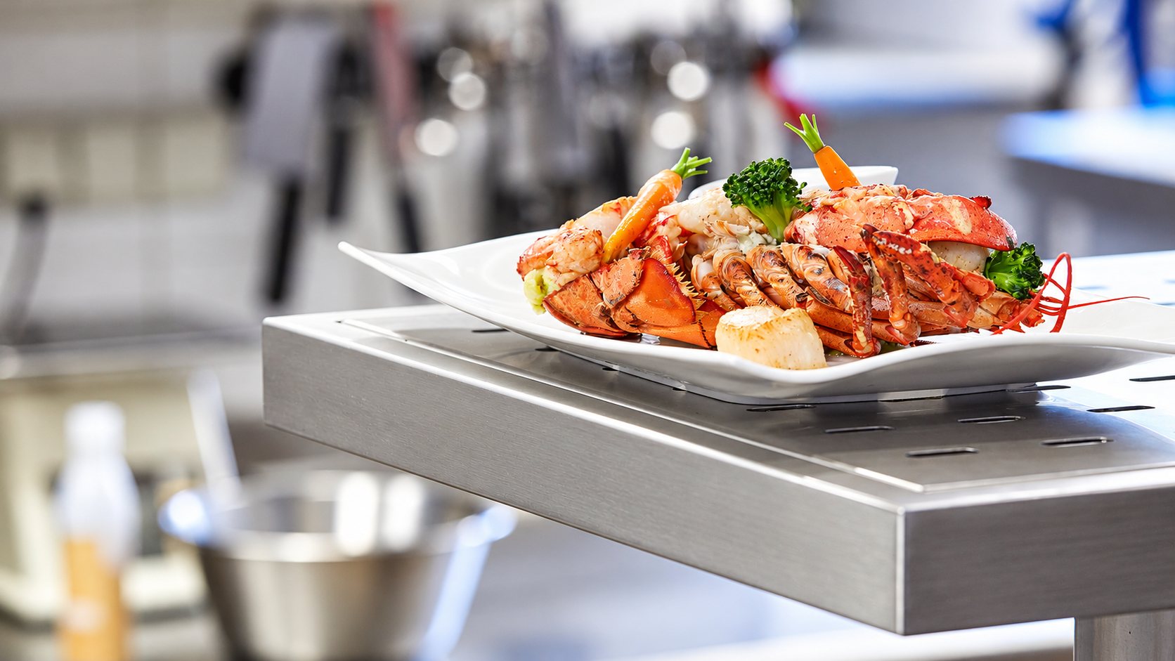Une assiette blanche garnie de homard, de Saint-Jacques, de brocoli et de petites carottes est posée sur un plan en inox dans une cuisine professionnelle.