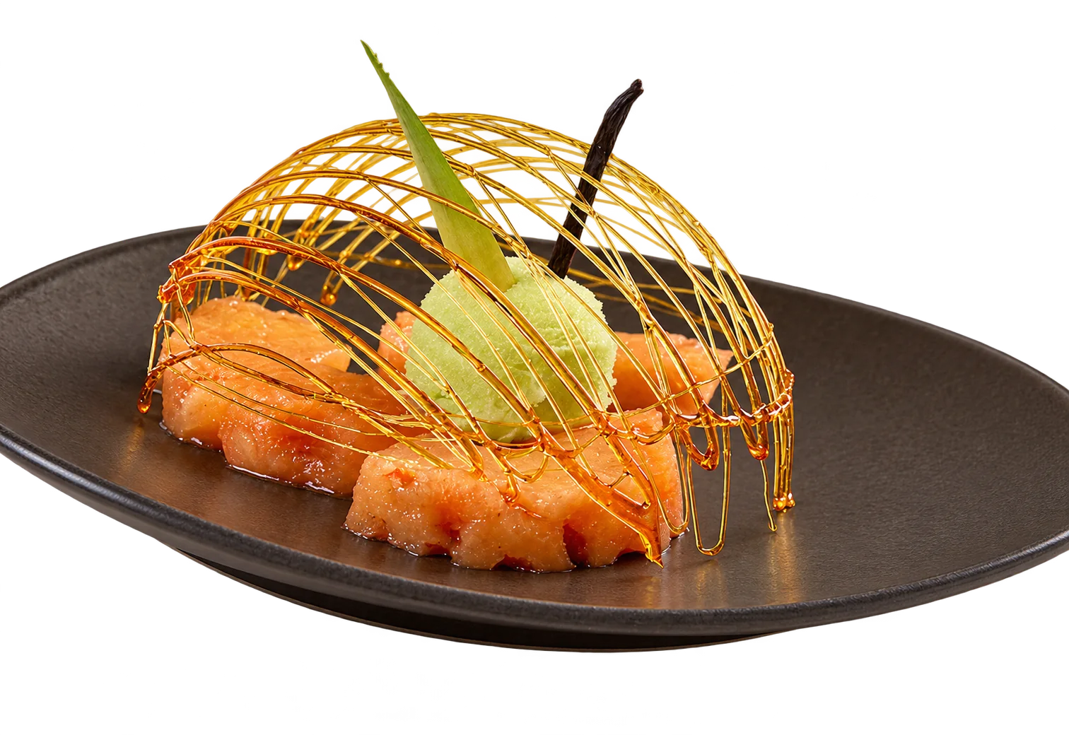 Une assiette brune creuse présente des tranches d'ananas rôti, une boule de glace verte, une gousse de vanille et un dôme de sucre filé doré.