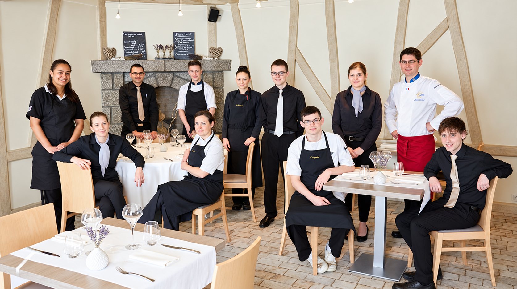 Onze membres de l'équipe d'un restaurant, en tenue noire et en veste de chef, posent autour de tables dressées devant une cheminée en pierre.