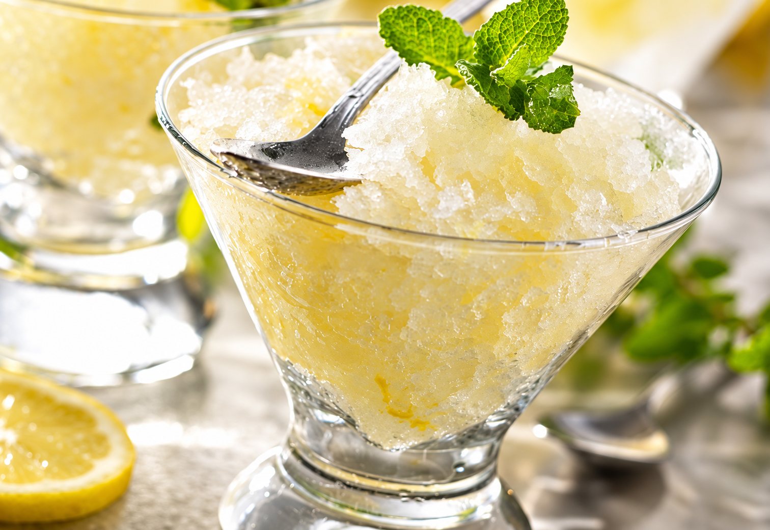 Une coupe en verre remplie de granité au citron, décorée d'une feuille de menthe fraîche et d'une cuillère, avec une tranche de citron au premier plan.