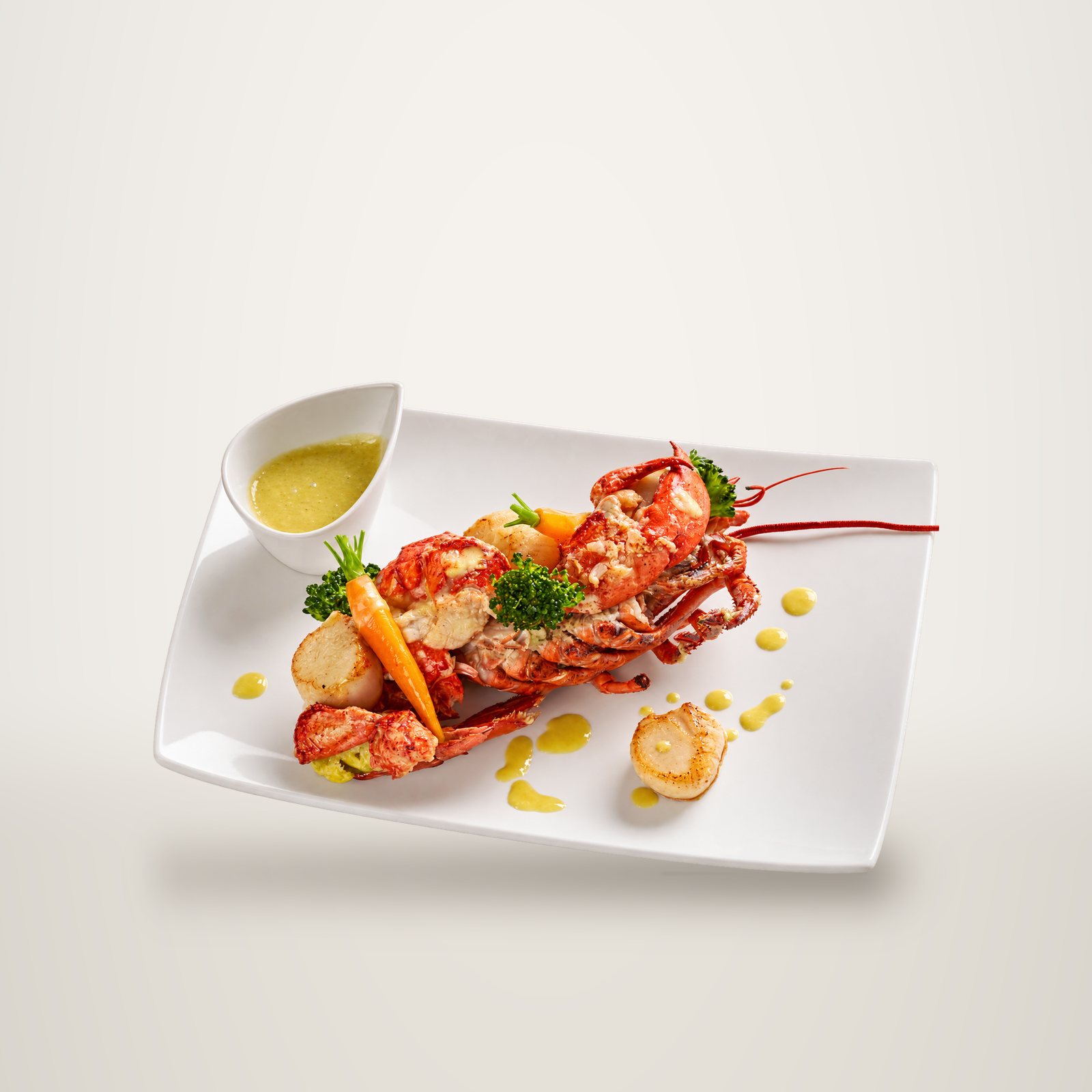 Assiette blanche garnie d'un homard grillé accompagné de noix de Saint-Jacques, de petites carottes, de brocolis et d'une sauce jaune servie dans un petit bol.