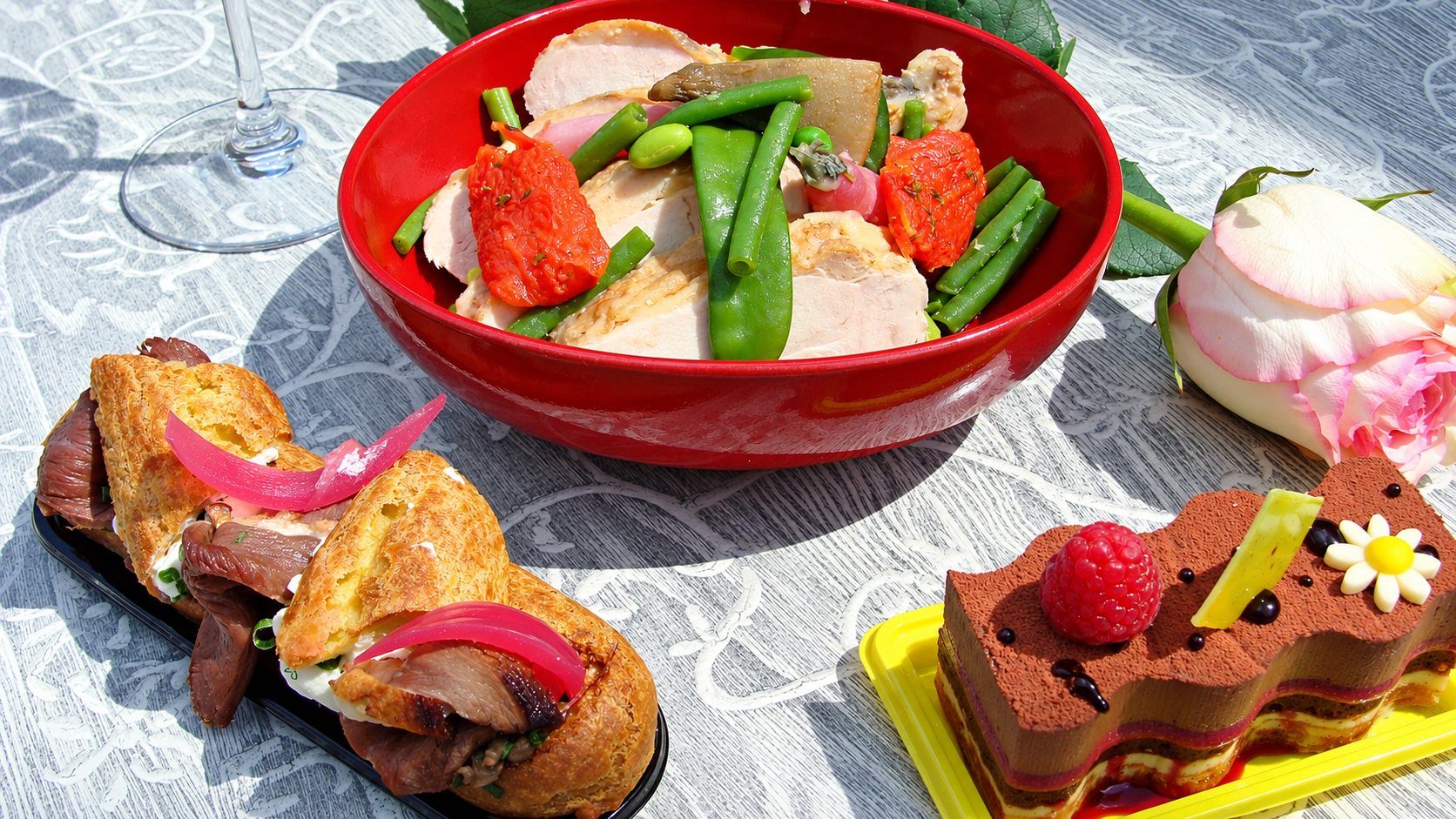 Sur une table extérieure, un saladier rouge de volaille aux haricots verts et tomates, un éclair salé, un gâteau au chocolat à la framboise et une rose rose.