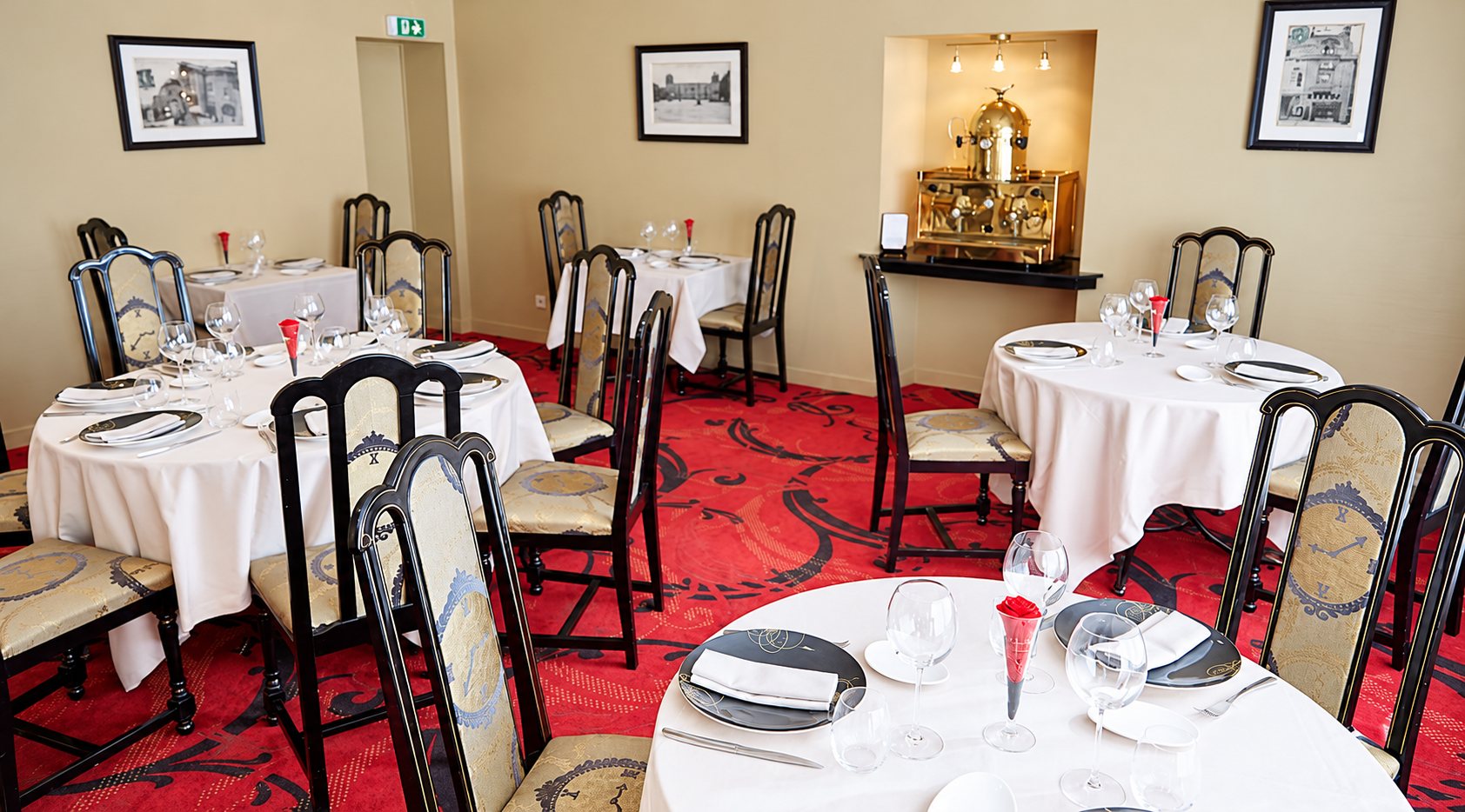Salle de restaurant aux tables rondes nappées de blanc et dressées, avec chaises noires et dorées, moquette rouge à motifs et machine à café dorée dans une niche.