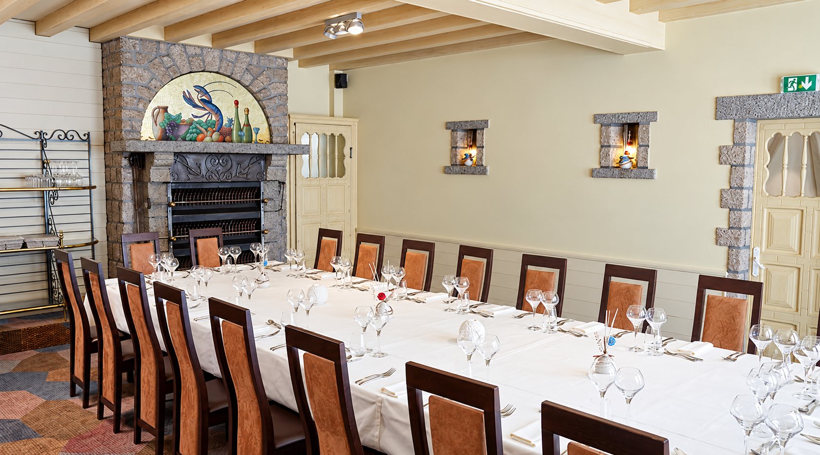 Une salle de restaurant aux murs clairs où une longue table à nappe blanche, entourée de chaises brunes, est dressée avec des verres devant une cheminée en pierre ornée d'un homard.