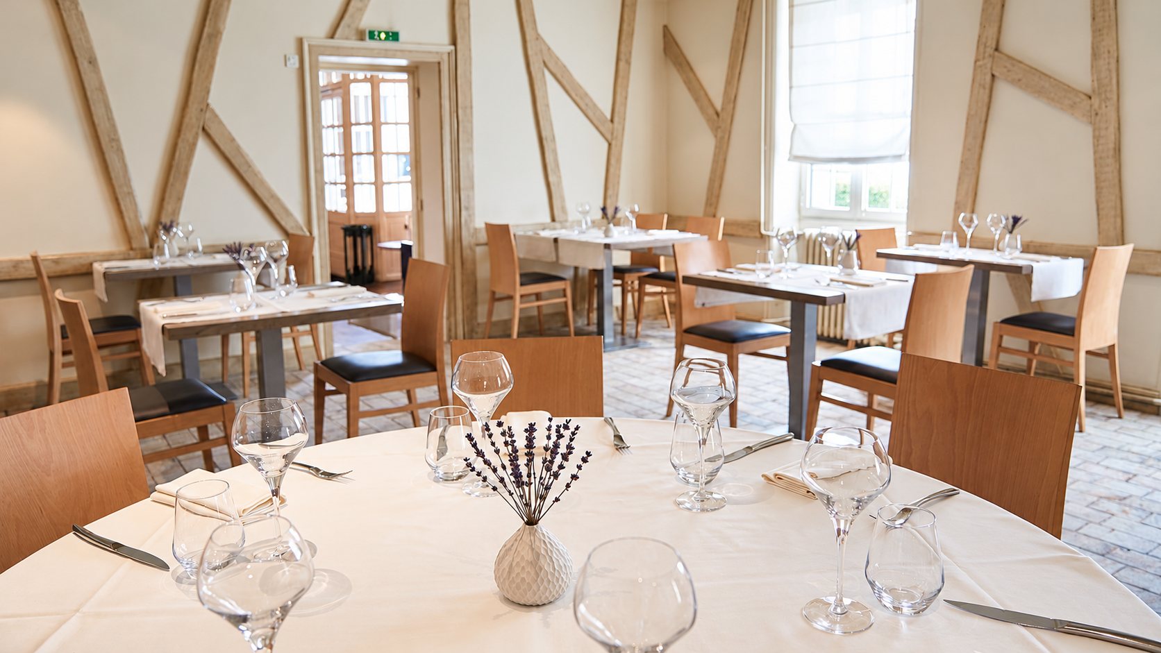 Salle de restaurant claire aux murs à colombages, avec des tables rondes dressées de verres, de couverts et d'un petit vase de lavande.
