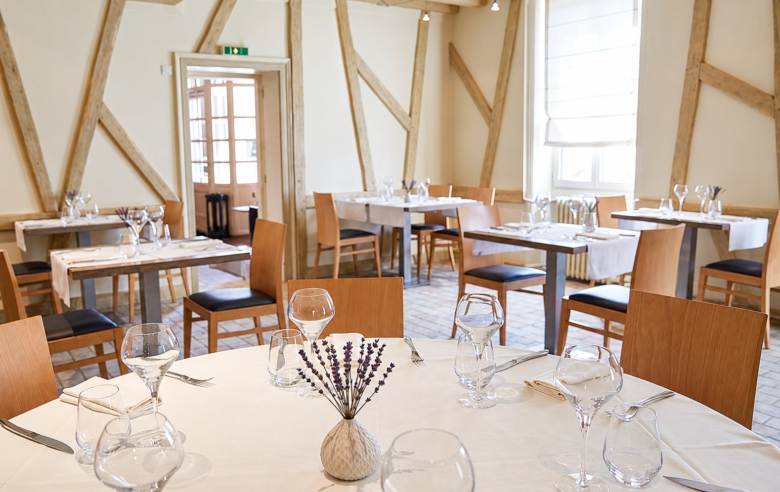 Salle de restaurant claire aux murs à colombages, avec des tables rondes et rectangulaires dressées de verres, de couverts et d'un petit vase de lavande.