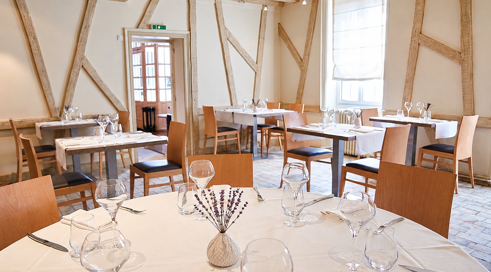 Salle de restaurant aux murs clairs à colombages, avec tables dressées de verres, couverts et un petit vase de lavande au premier plan.