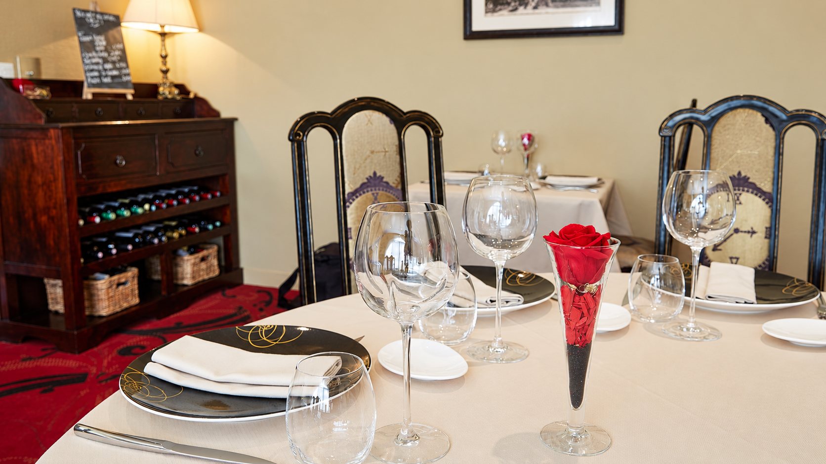 Table ronde élégamment dressée avec assiettes, verres et une rose rouge dans un vase, devant un meuble en bois sombre contenant des bouteilles de vin.